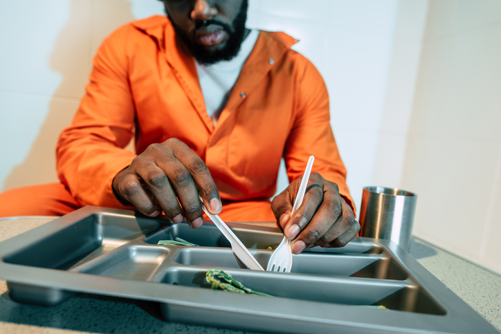An incarcerated person eats prison food from a tray with a plastic knife and fork.